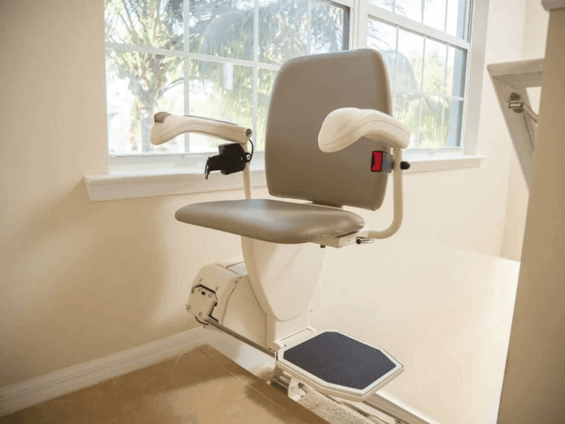 The Harmar stair lift, which features a light grey plastic seat, stops at the top of wooden stairs. Trees are visible outside the window behind it