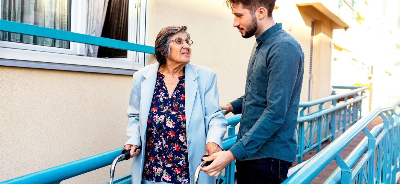A senior woman using a walker is outside walking with her younger male caregiver.