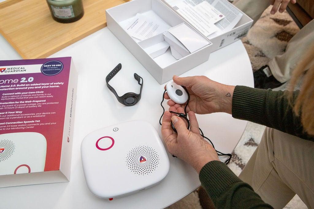 A Medical Guardian Home 2.0 box and related devices on a table, hands holding the medical alert pendant