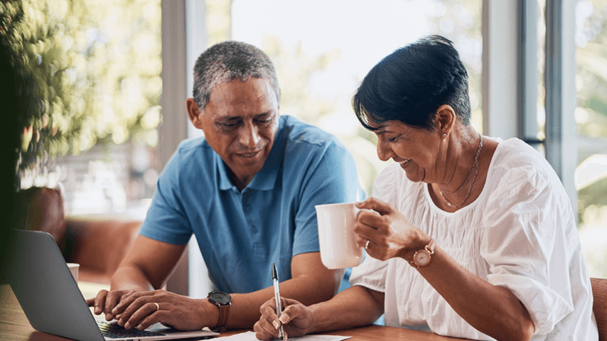 An older couple sitting at a table with a laptop, signing a document