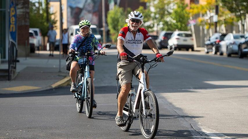 Two people riding bicycles on a street smile at the camera.