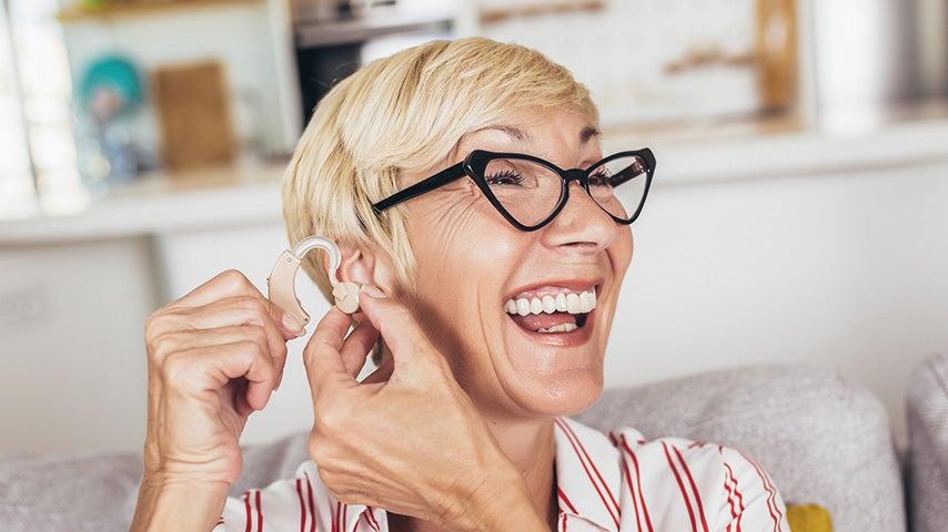 smiling older woman with short blonde hair putting hearing aid in left ear