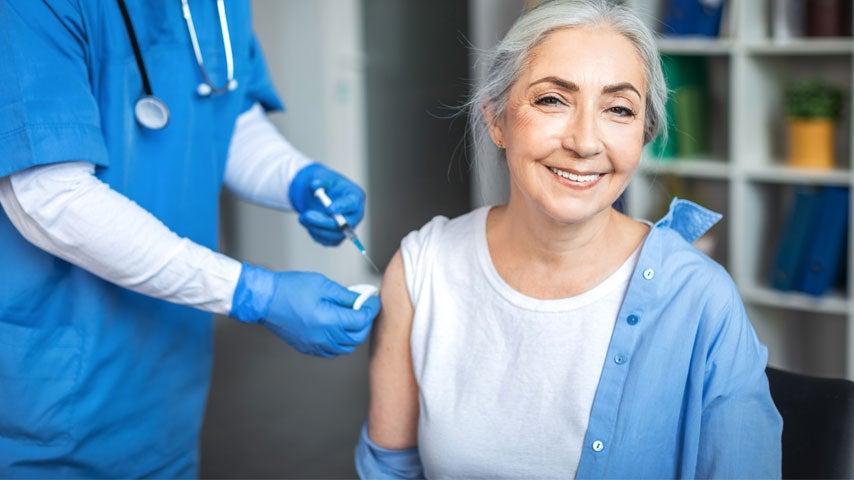 An older adult woman with gray hair is smiling while getting an mRNA covid vaccine from her doctor.