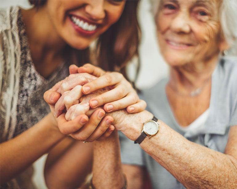 A younger female caregiver holds the hands of a senior Caucasian woman who is smiling.