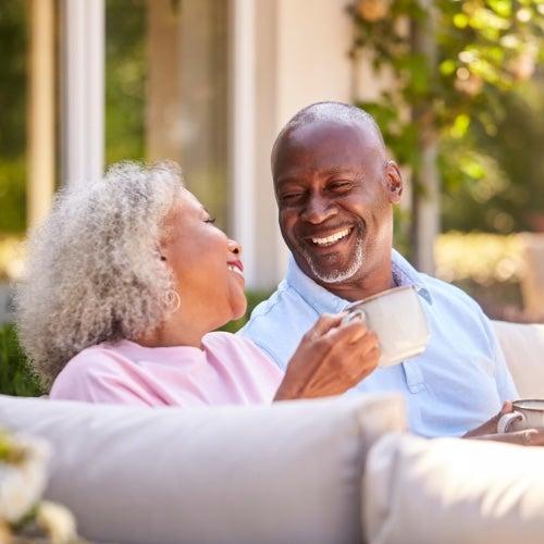 Two older adults sit on an outdoor couch and laugh while drinking coffee