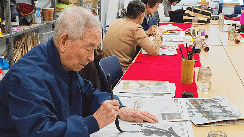 older Asian man painting Chinese characters onto a newspaper during art class