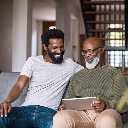Two men sit on a couch and smile while looking at a tablet the older man is holding
