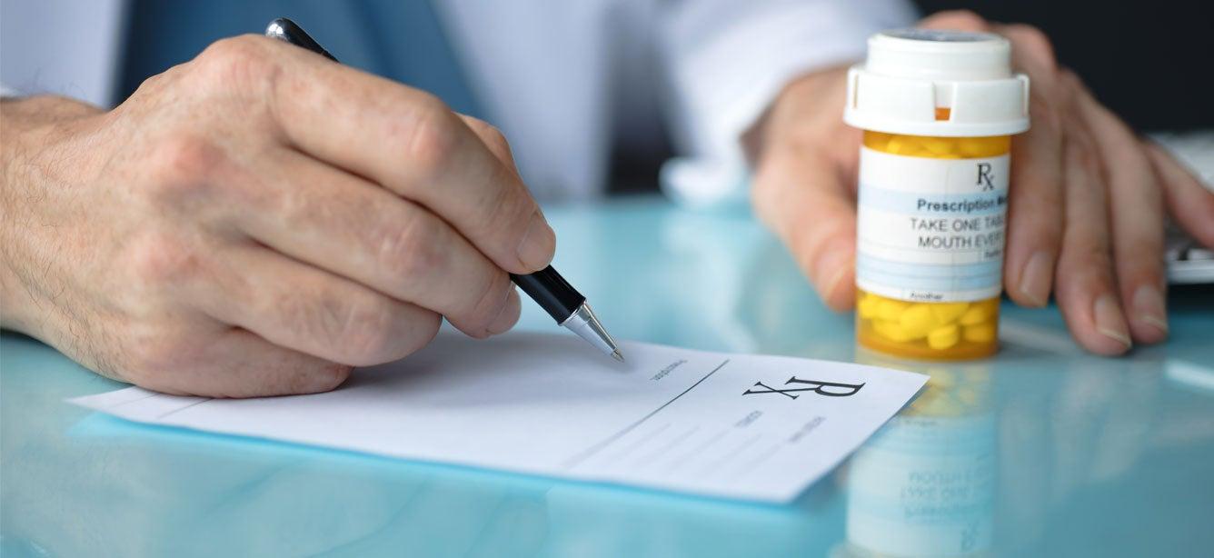A hand writing on a prescription pad, next to a yellow pill bottle with a label and instructions, on a glass surface.