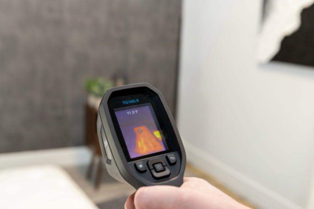 A person holds a heat sensing gun, which shows oranges and yellows on the screen