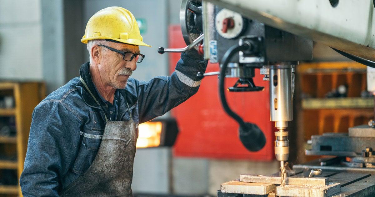 A senior man with a yellow hard hat and glasses works with a large drill bit at a plant workshop.