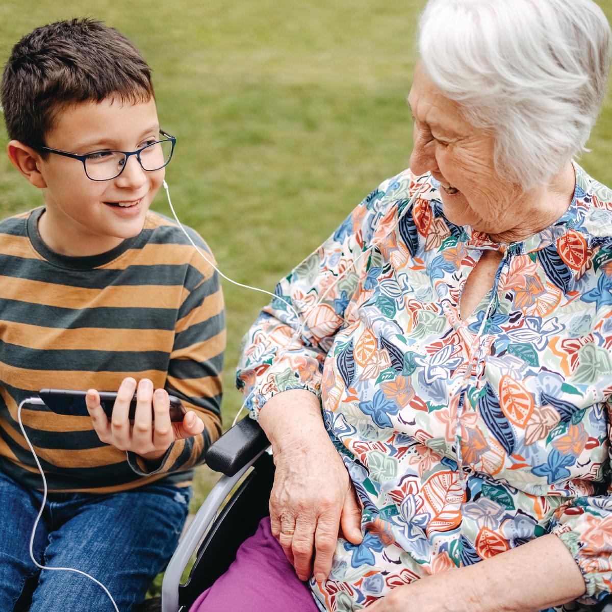 An older woman is sitting with a young boy, and they're listening to music on his mobile device, sharing earphones so both can listen to the music.