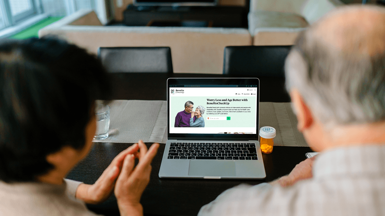 An older couple sits at a dining table, viewing Benefits CheckUp on a laptop.