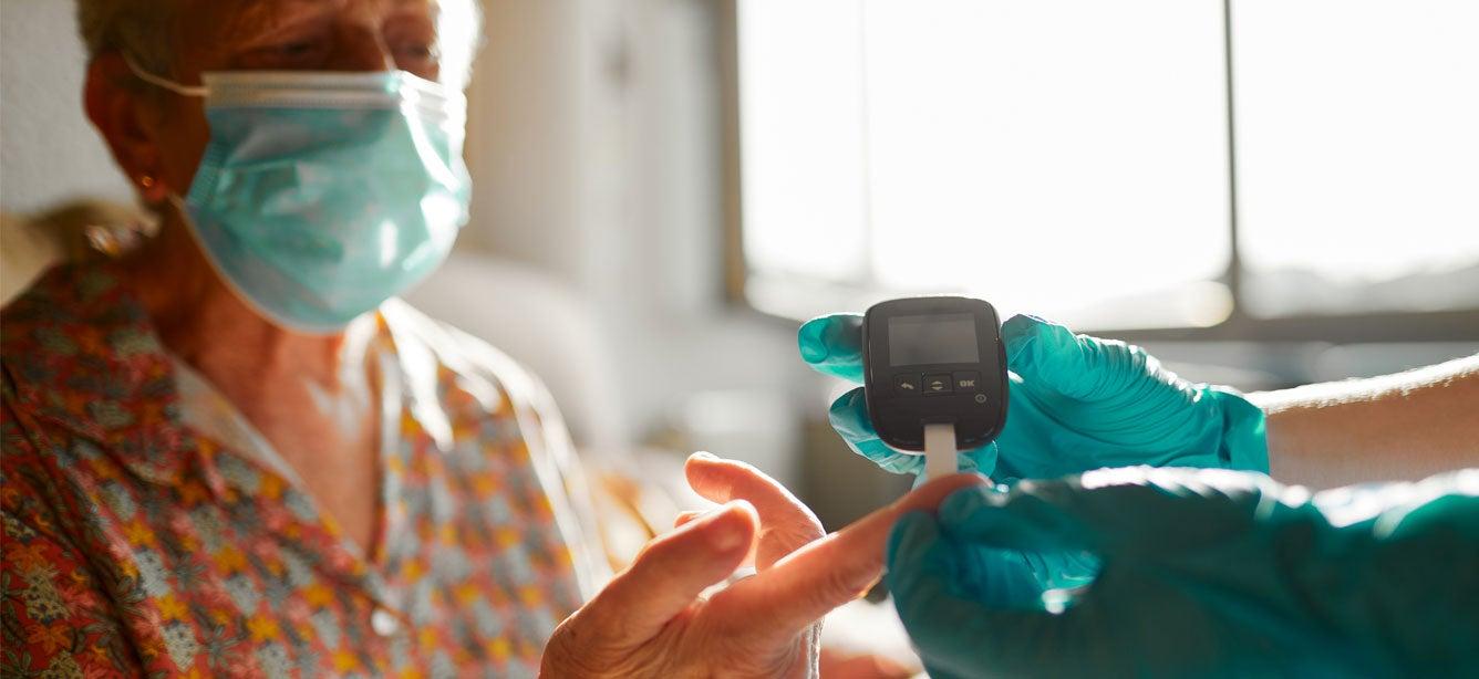 A senior woman with diabetes is wearing a mask and getting her insulin checked by a nurse wearing blue latex gloves.